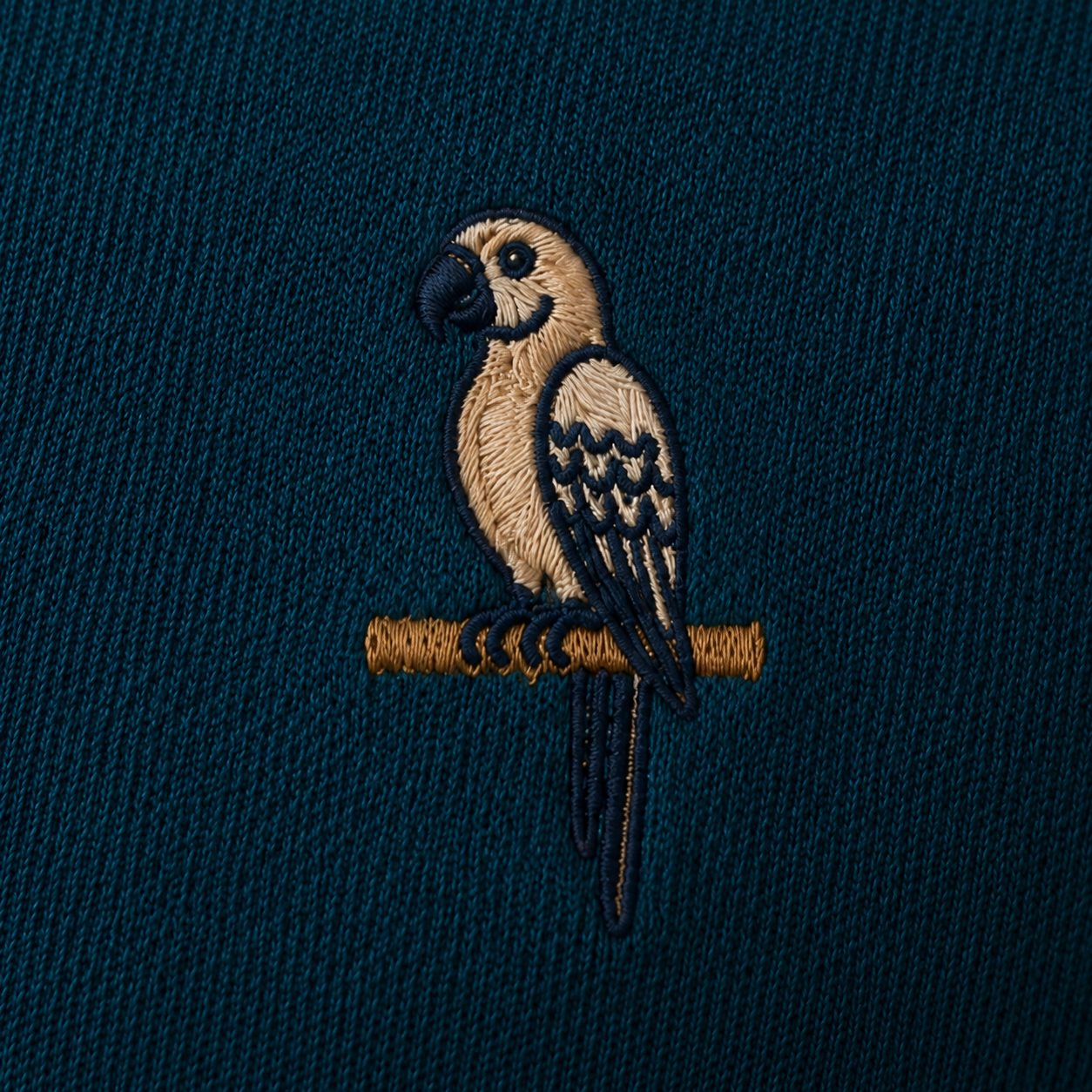 Tonal chest embroidery of a parrot perched on a brass bar, cream and brass thread on deep teal piqué fabric