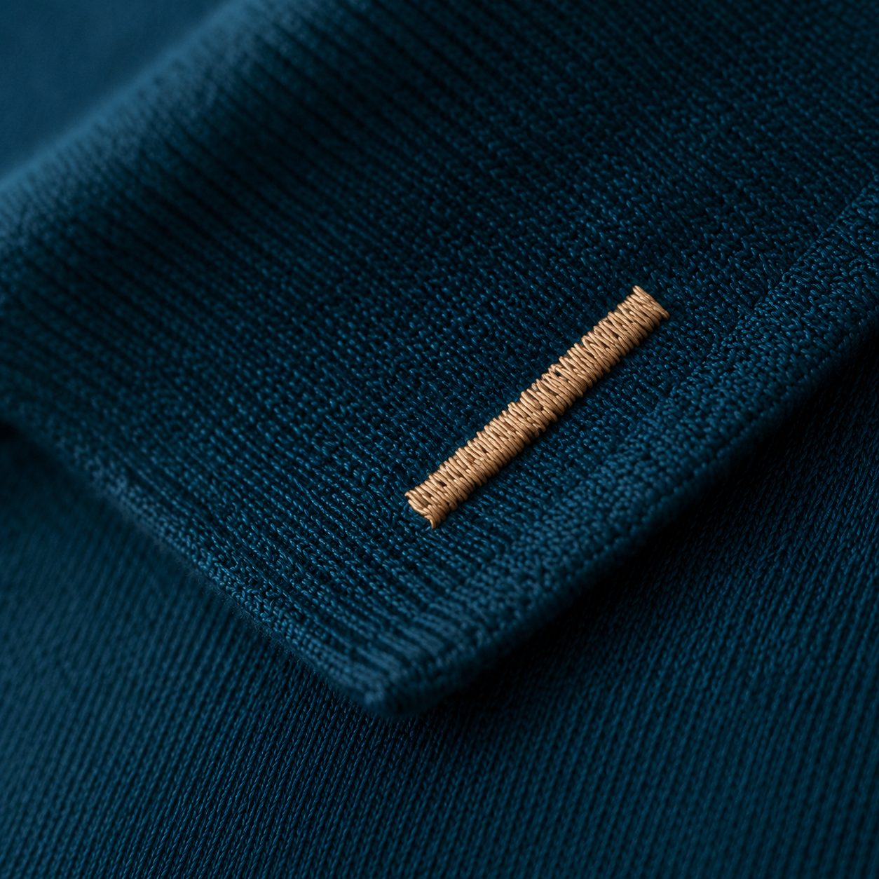 Close-up of the sleeve cuff showing a single vertical brass perch-bar embroidery on the ribbed cuff edge